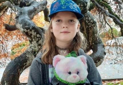 A blond child holding a plush animal standing in front of a tree