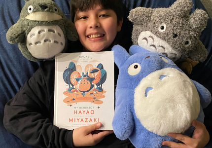 A boy laying down on a bean bag chair holding a book with 3 Totoro toys around him